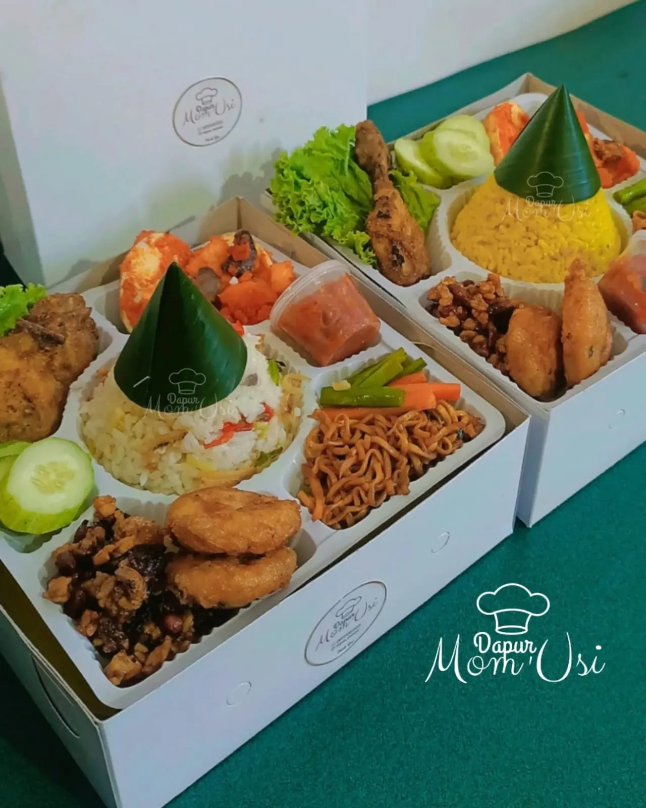 Dua nasi box terbuka berisi nasi kuning dan nasi putih dengan ayam goreng, mi goreng, orek tempe, dan sambal