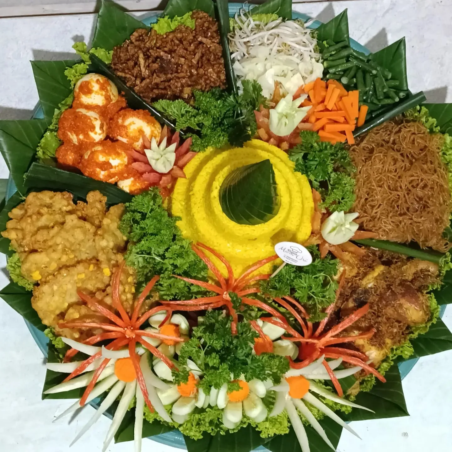 Tumpeng tampah tampak atas dengan nasi kuning di tengah dikelilingi orek tempe, telur balado, ayam goreng, bihun, dan hiasan ukir sayuran